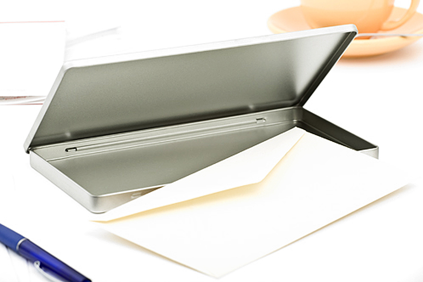 Boîte en aluminium ouverte au format DIN-Lang avec couvercle à charnière déplié, une enveloppe partiellement posée dedans, arrangée sur un bureau avec une tasse et un stylo en arrière-plan.