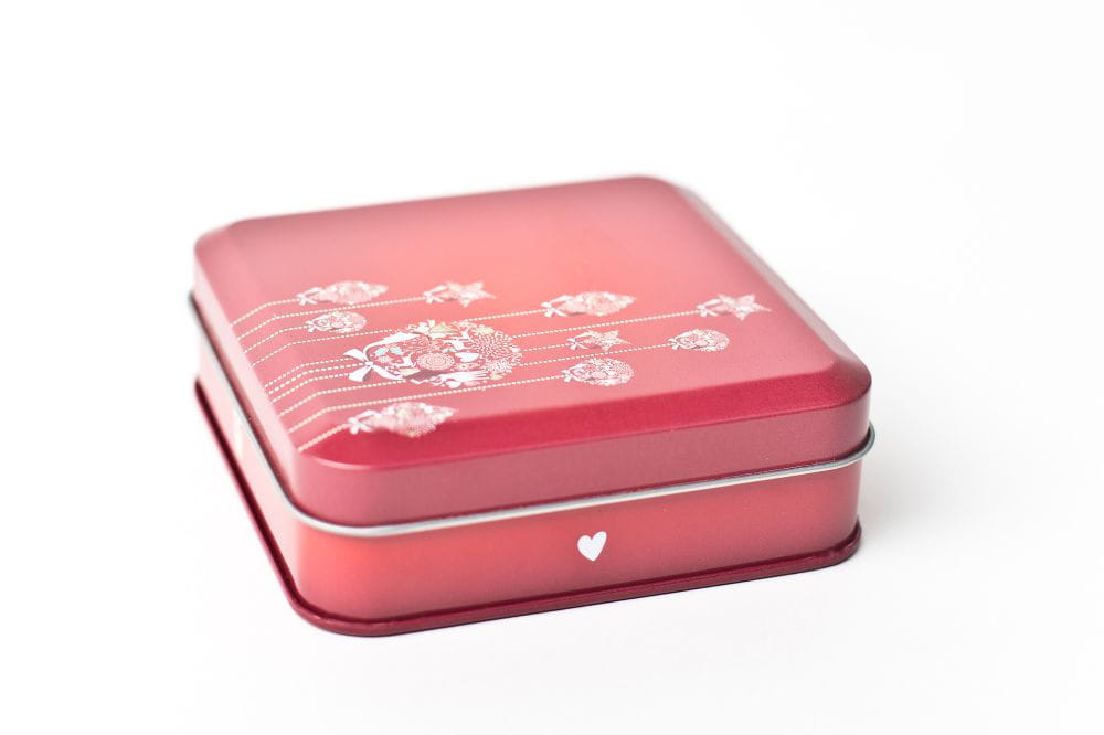 Boîte métallique rectangulaire fermée en rouge avec ornements floraux et motifs de cœurs sur fond blanc.